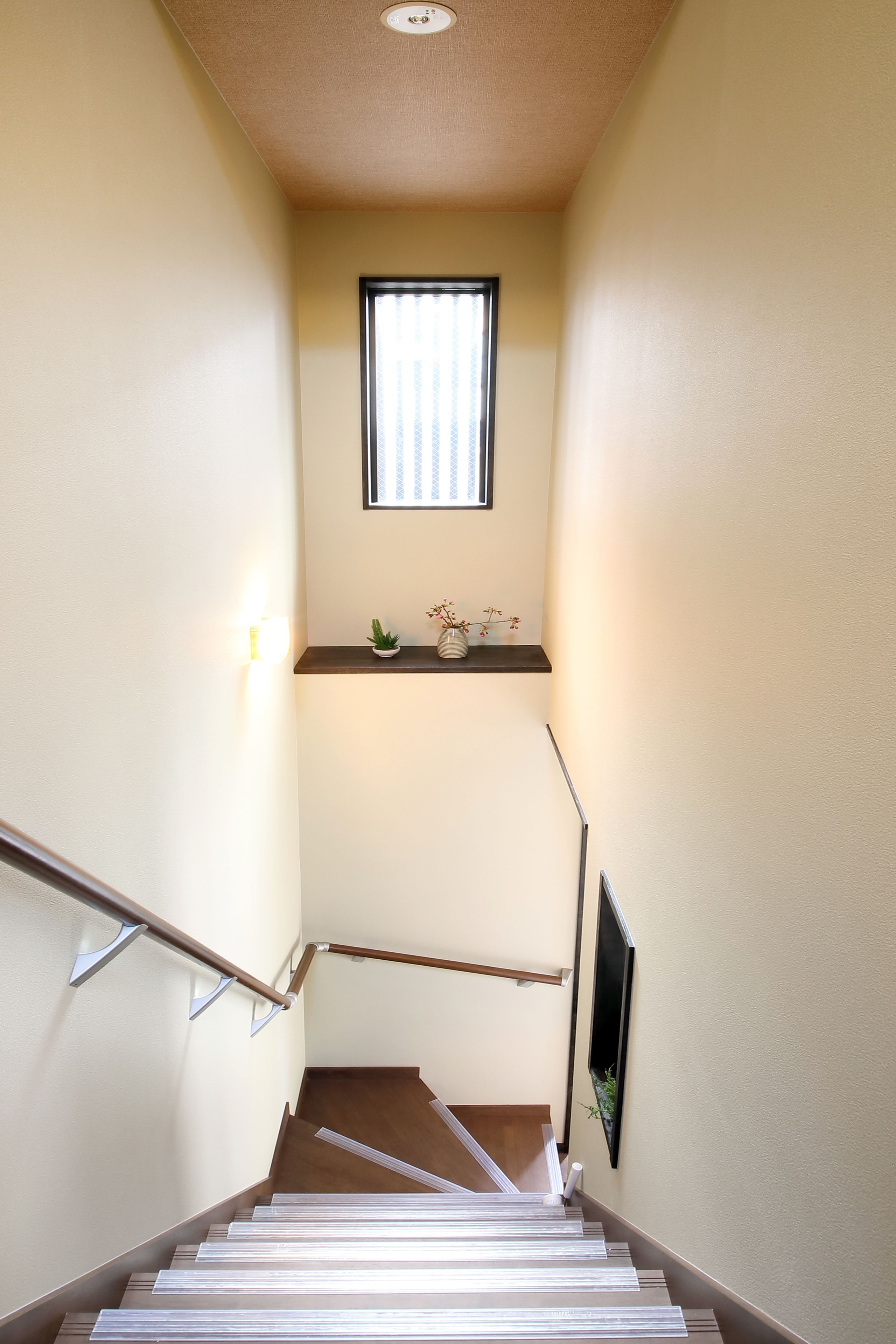 Un escalier avec une rampe, une petite étagère avec une plante et une fenêtre avec des stores.