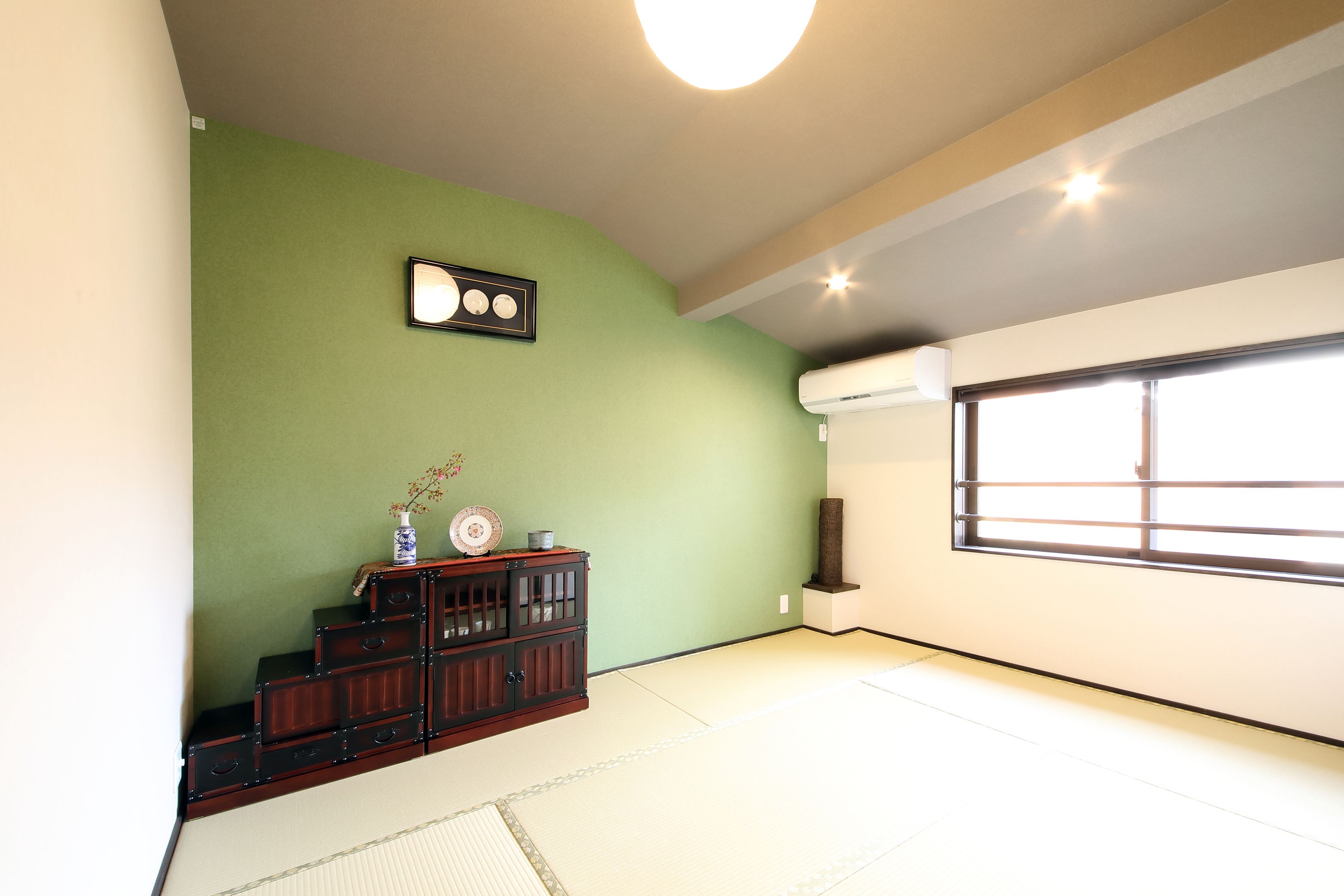 A room with tatami flooring, a green wall, and a traditional Japanese cabinet with a vase.