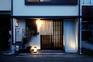 Un bâtiment moderne doté d’une entrée sécurisée par un portail, d’une enseigne indiquant « Résidence d’invités » et d’un petit espace extérieur agrémenté d’un pot de fleurs et d’un luminaire.
