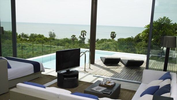 Habitación de hotel moderna con un balcón grande, televisor, ordenador portátil y zona de comedor con vistas al mar.
