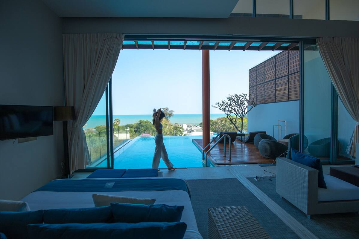 A modern hotel room with a large bed, a TV, and a view of a swimming pool and ocean.