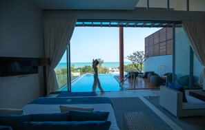 Quarto de hotel moderno com uma cama grande, televisão e vista para uma piscina e oceano.