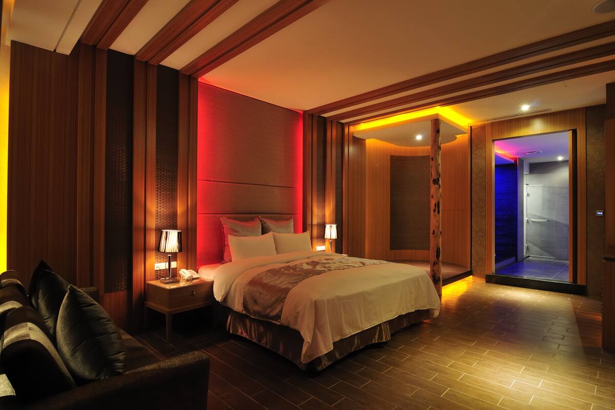 Ein modernes Hotelzimmer mit einem großen Bett, zwei Nachttischen mit Lampen, einem dunkelbraunen Ledersofa und Holzböden.