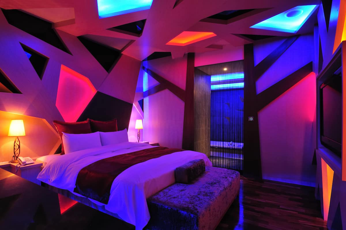 Ein modernes Hotelzimmer mit einem großen Bett, einem Nachttisch, einer Lampe und Blick durch Vorhänge auf die Außenwelt.
