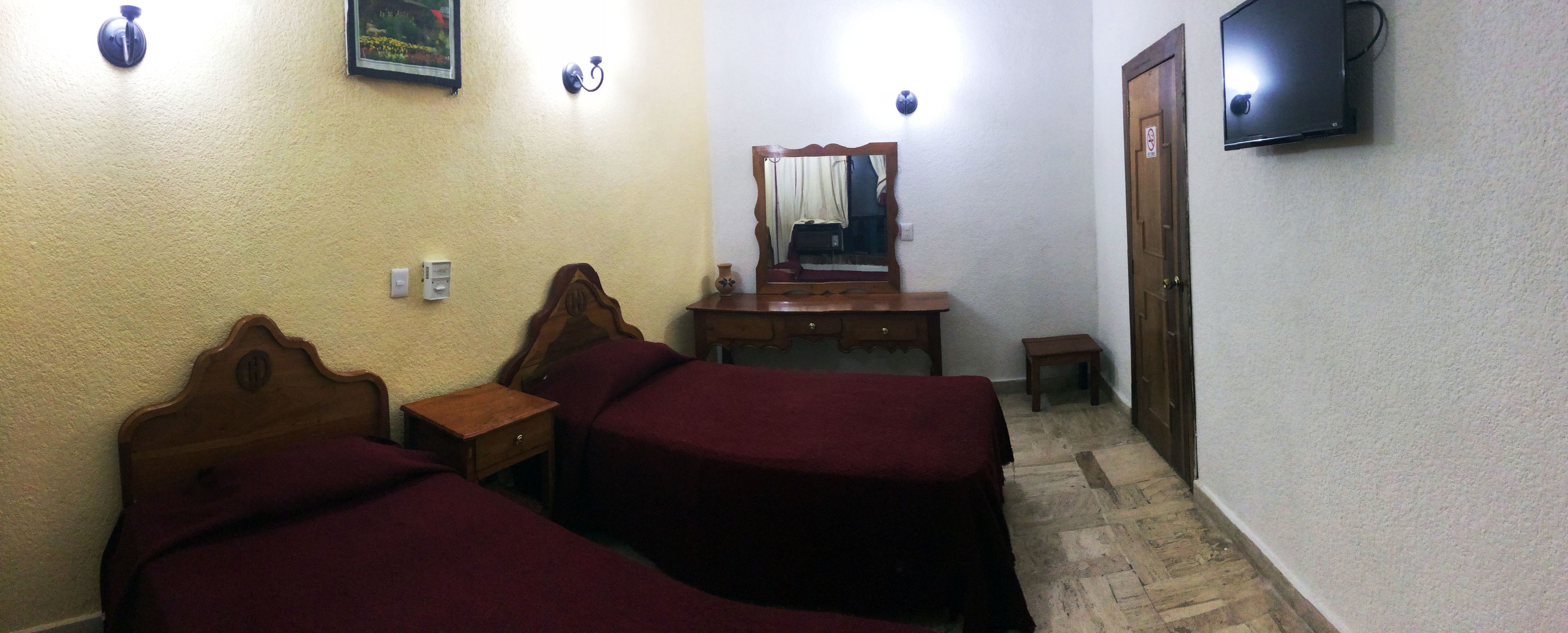 A small, dimly lit room with two wooden beds, a wooden dresser, a mirror, a television, and a small table.