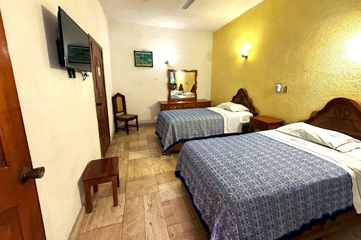 Une chambre d’hôtel avec deux lits, une télévision, un ventilateur de plafond, une chaise en bois, un bureau et un miroir.