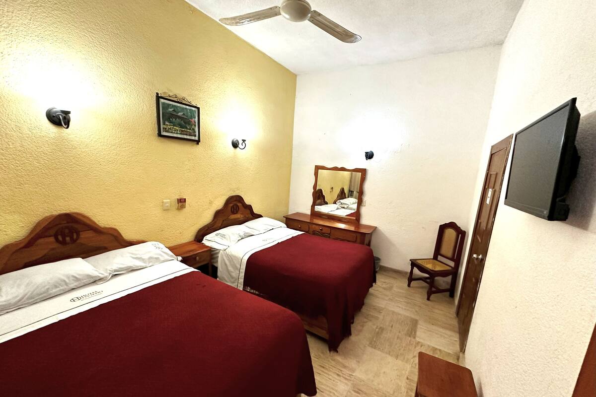 Une chambre d’hôtel avec deux lits, une commode en bois, une chaise, un téléviseur fixé au mur et un ventilateur de plafond.