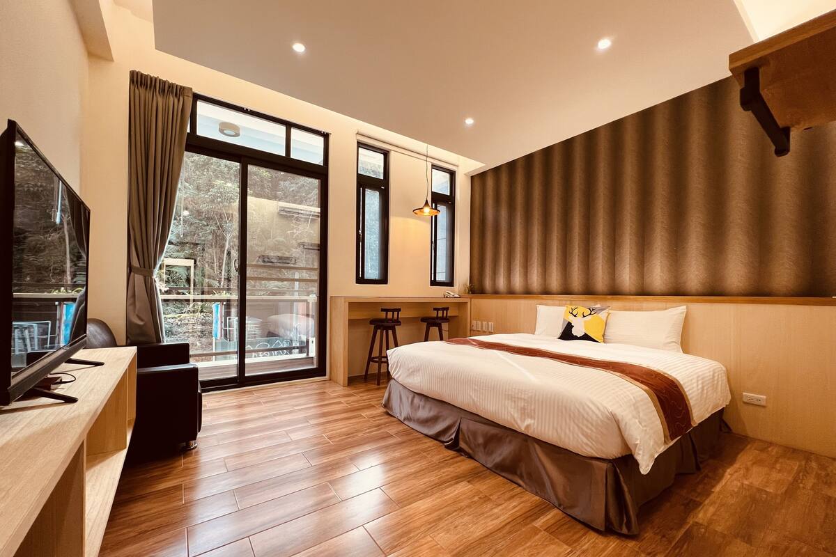A modern hotel room with a large bed, a desk with a computer, and a balcony with a view of trees.