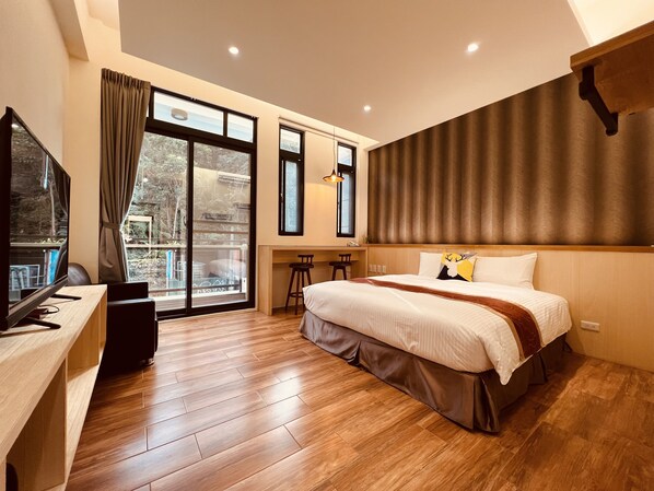 A modern hotel room with a large bed, a desk with a computer, and a balcony with a view of trees.