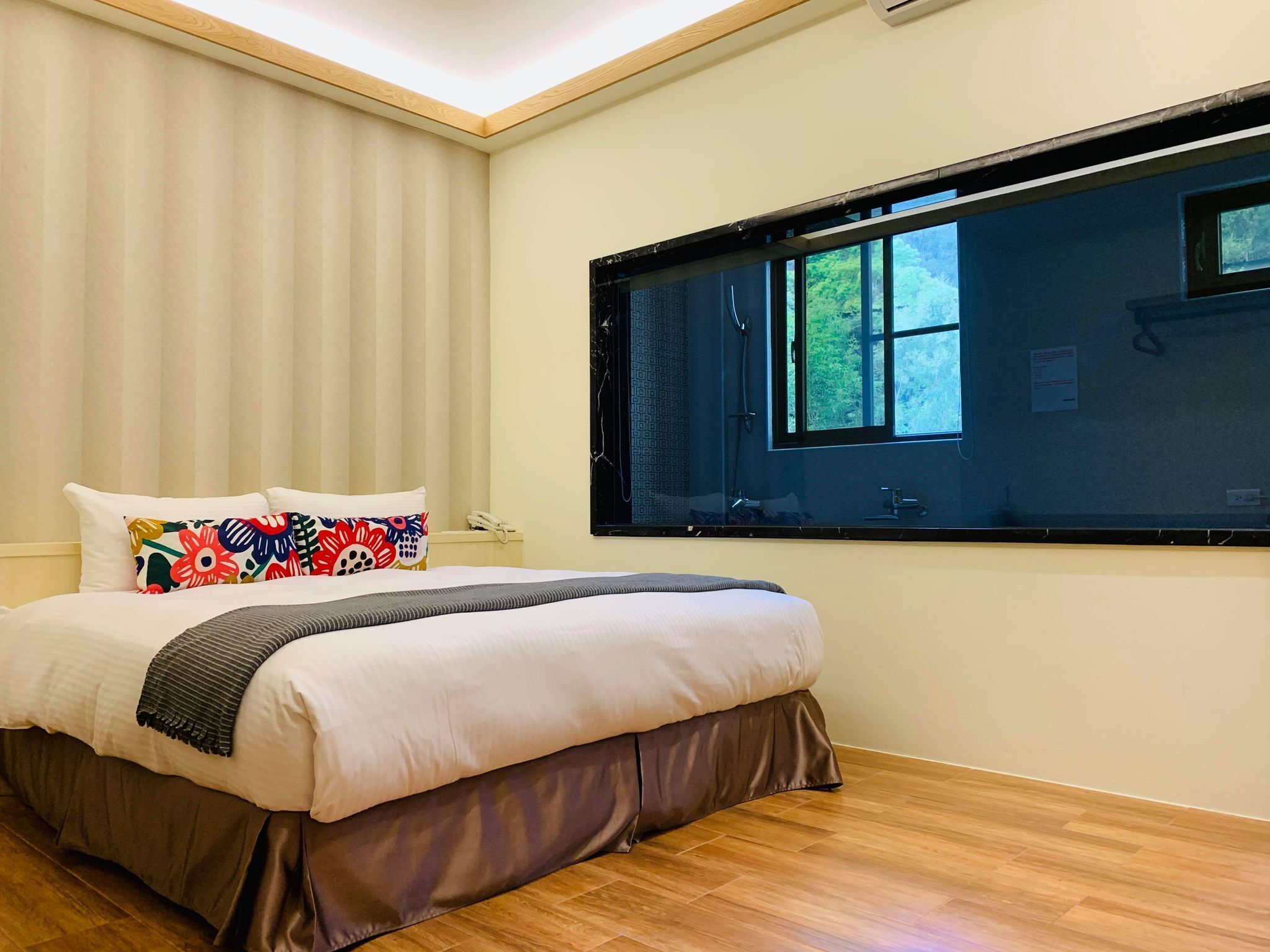 A bedroom with a large bed, a window with a view of greenery, and a wooden floor.