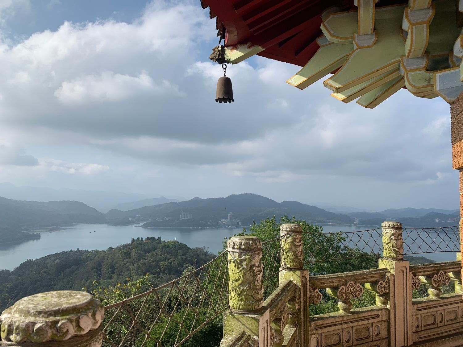 Vista desde un lugar elevado que domina un lago y montañas, con una estructura arquitectónica tradicional que presenta barandales ornamentales y una campana.