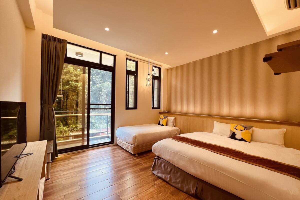 A hotel room with two beds, a TV, and a balcony with a view of greenery.