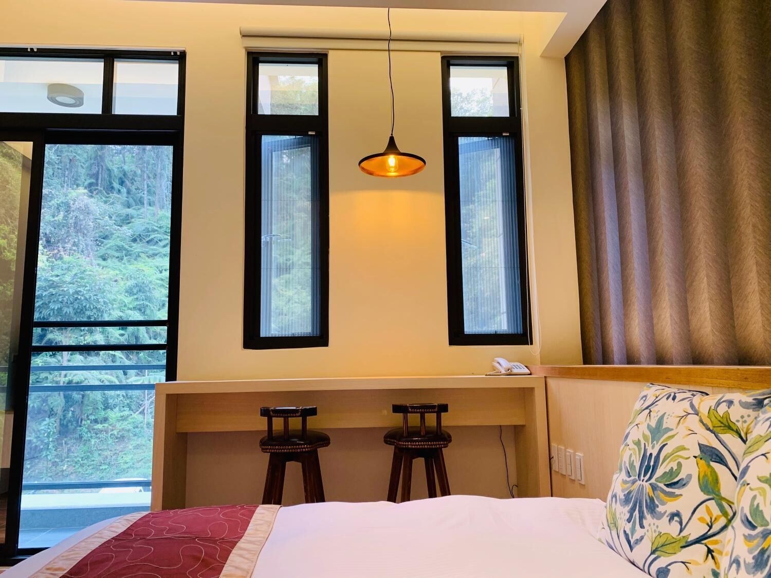 A bedroom with a bed, two stools, a desk, and a hanging light fixture.