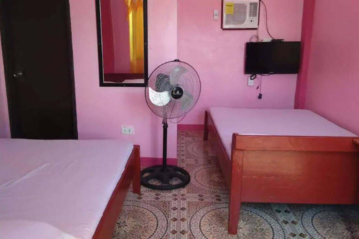 Une chambre avec deux lits en bois, un ventilateur sur pied, un climatiseur fixé au mur et un miroir.