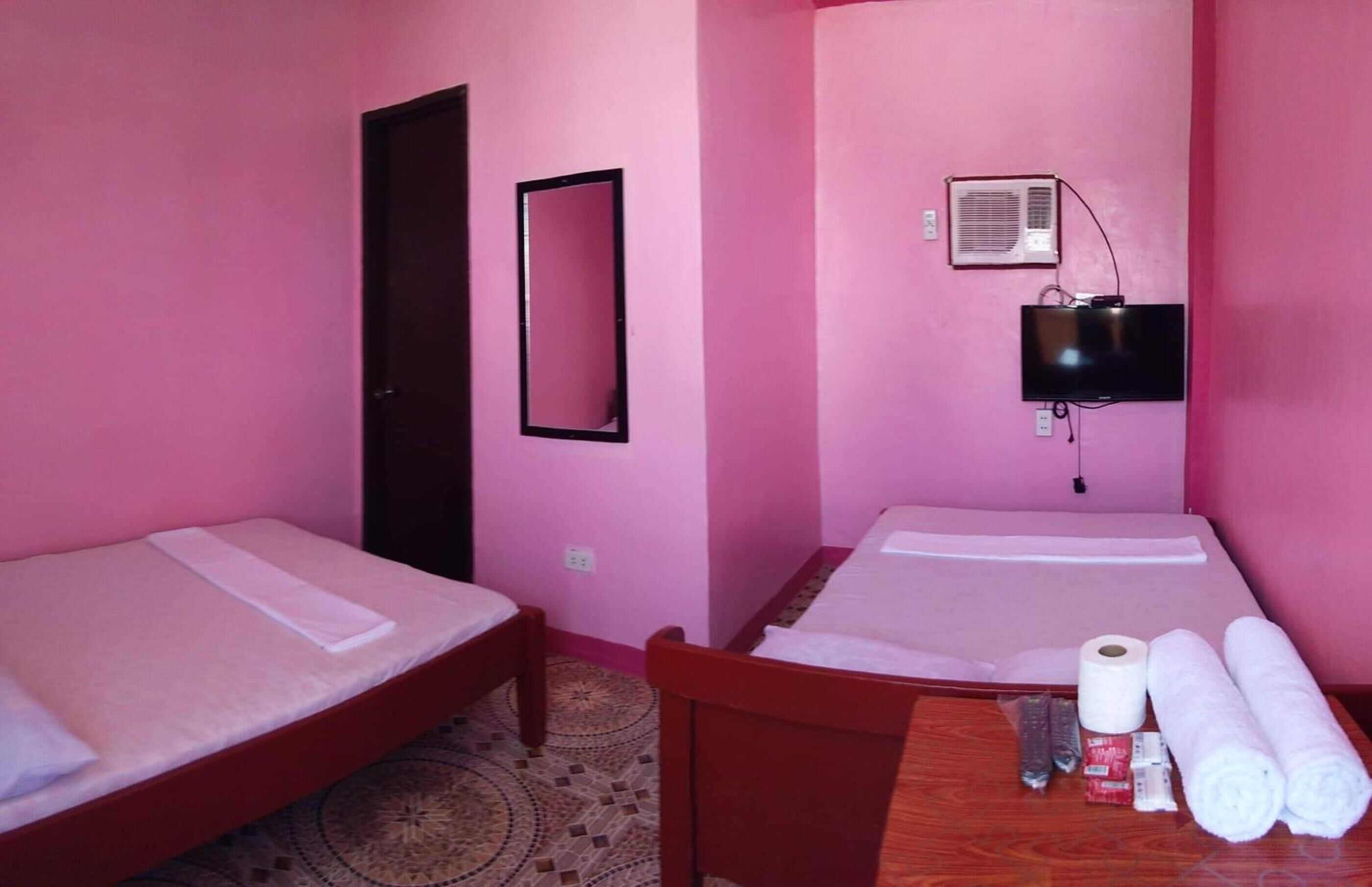 Quarto com duas camas de solteiro, uma cadeira de madeira, uma mesa com artigos de higiene pessoal, um espelho e um ar condicionado fixado na parede.