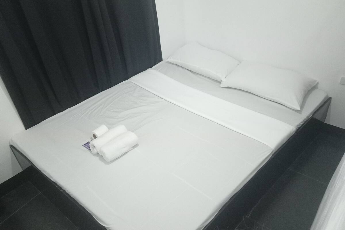 Une petite chambre avec un lit simple, recouverte d’une courtepointe blanche unie, une serviette blanche soigneusement pliée et un rideau noir sur le côté gauche.