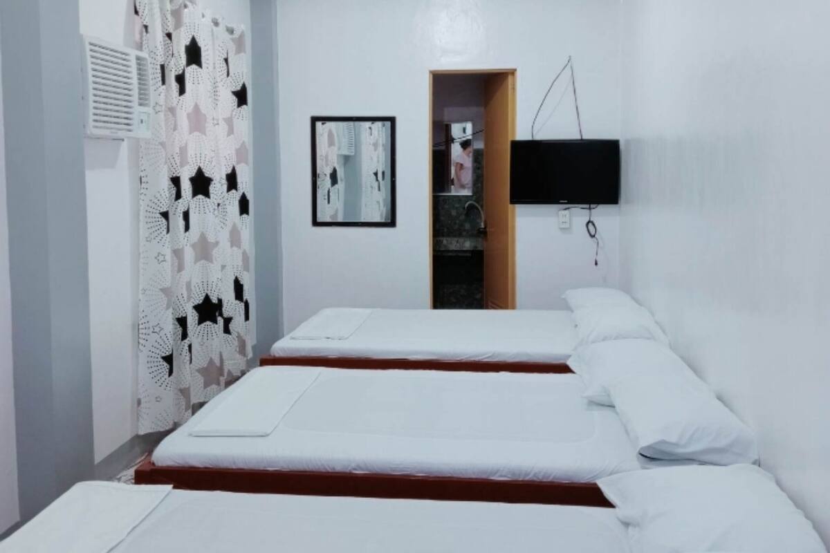 Une chambre avec trois lits alignés en rangée, un téléviseur à écran plat fixé au mur et une porte donnant sur une salle de bain.