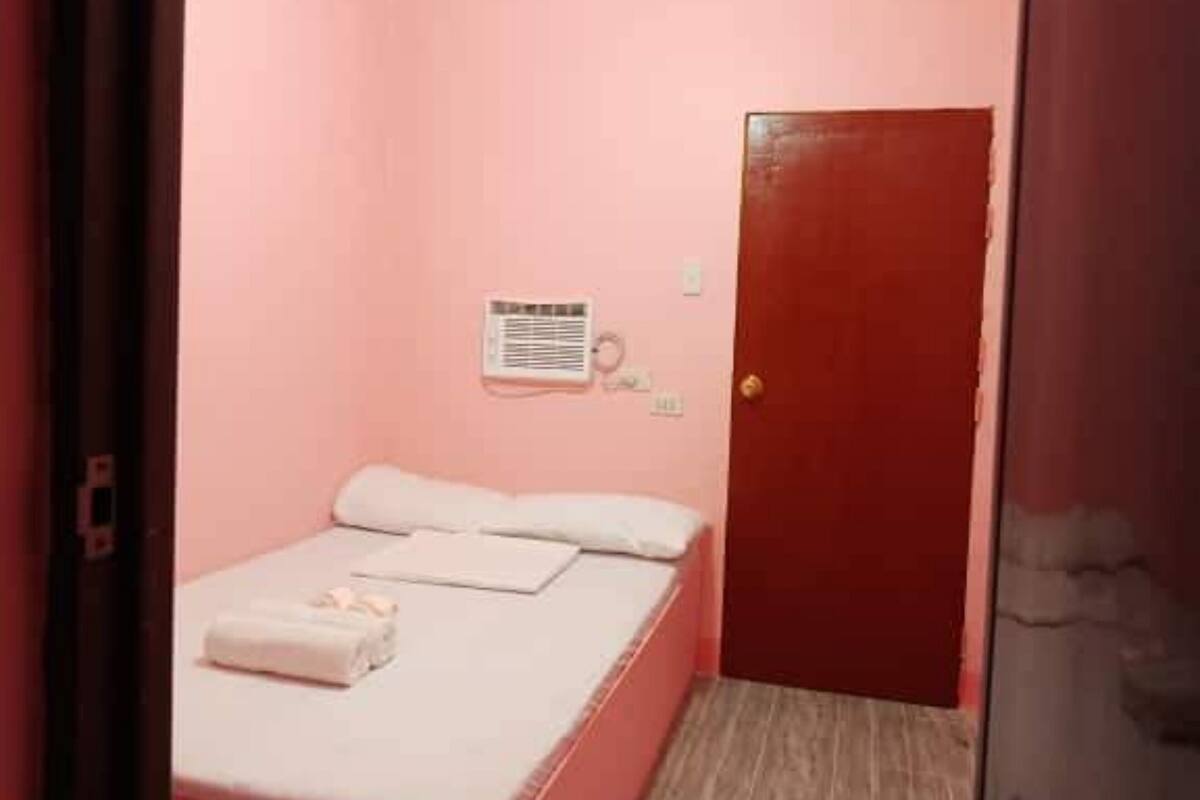 Une petite pièce aux murs roses, avec un lit simple, une porte et un climatiseur fixé au mur.