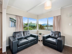 A room with two black leather sofas, a small table with a bowl, and large windows offering a view of greenery and mountains. - Taupo Panorama - Lake Taupo Holiday Home (Taupo Central)