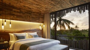 Slaapkamer met een houten plafond, een groot bed met witte lakens, een bankje en uitzicht op palmbomen en een zonsondergang.
