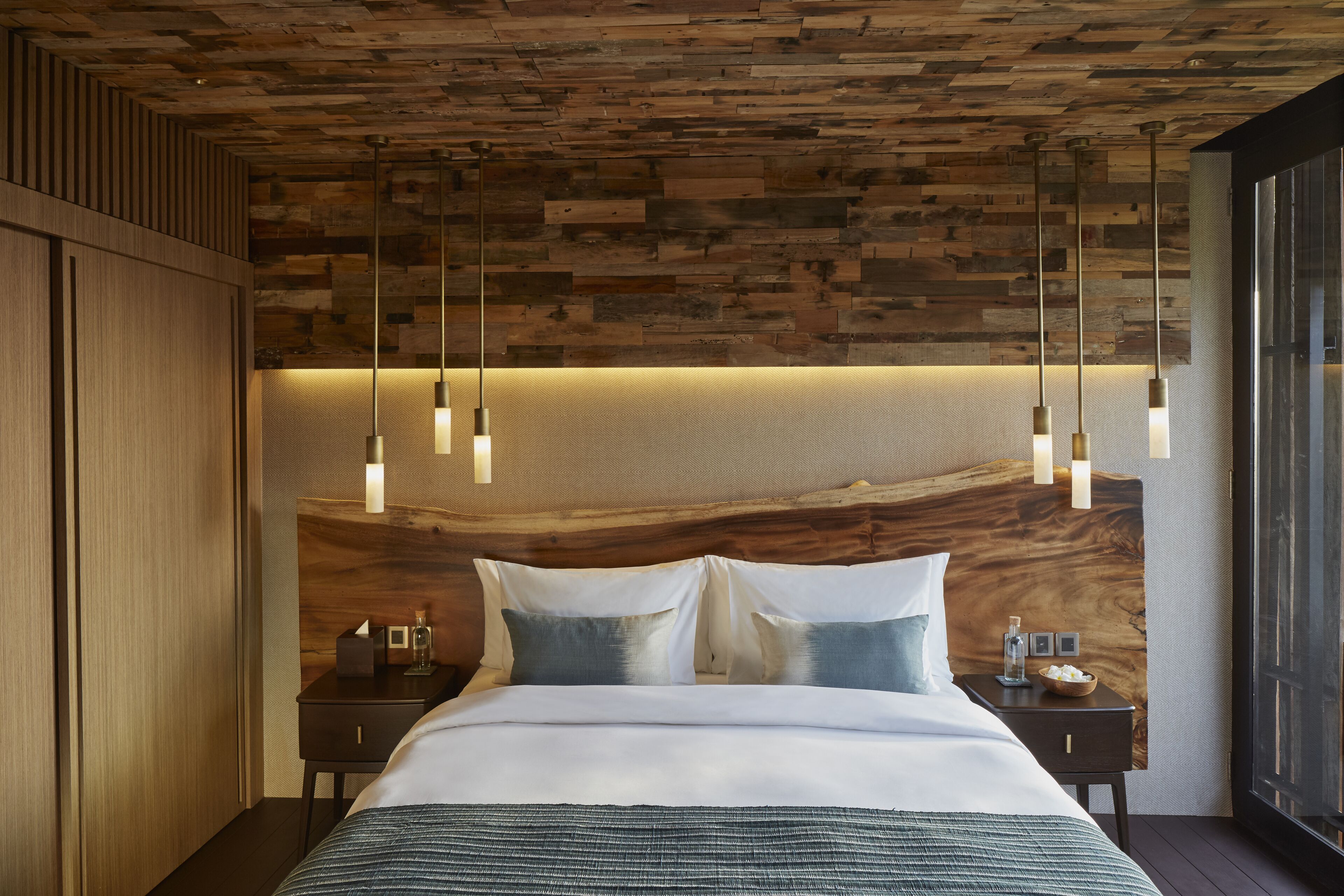 A modern bedroom with a wooden headboard, a large bed with white linens, a nightstand with a lamp, and a view of the outdoors through a window.