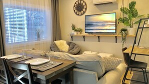 A modern living room with a sofa, a dining table with chairs, a TV mounted on the wall, and a window with blinds.