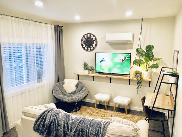 A living room with a sofa, a grey pet bed, a television, a clock, and a plant shelf. - Private & Comfortable Guesthouse close to Portland +AC! (Vancouver)