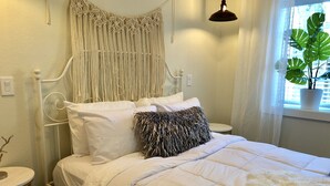 A bed with white bedding, a macrame headboard, a round bedside table, and a potted plant by the window.