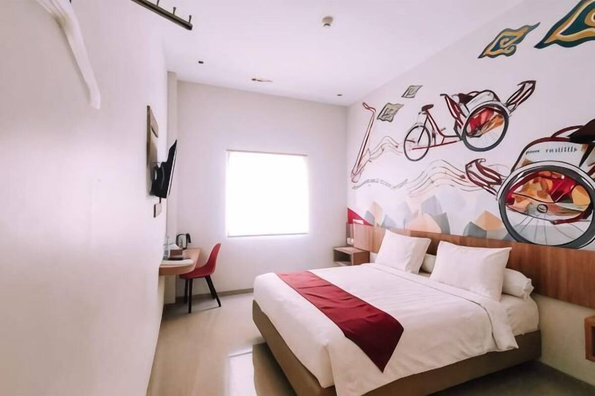A hotel room with a bed, a desk, a chair, a window, and a wall with bicycle illustrations.