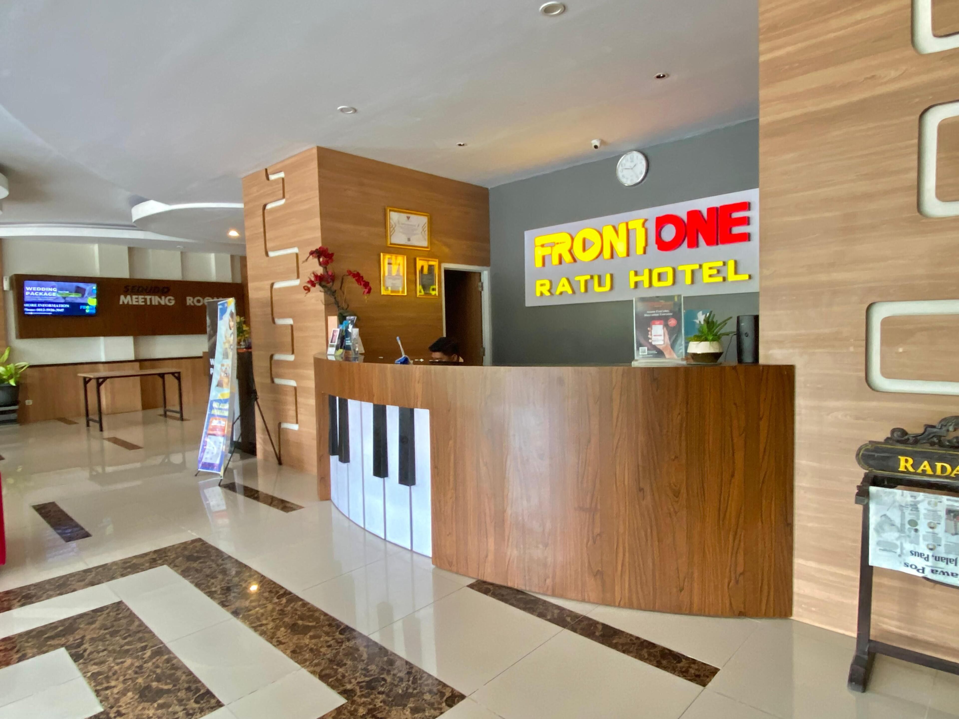 Un espace d’accueil d’hôtel avec un comptoir de réception en bois, un écran numérique et une enseigne indiquant « FRONONE Ratu Hotel ».