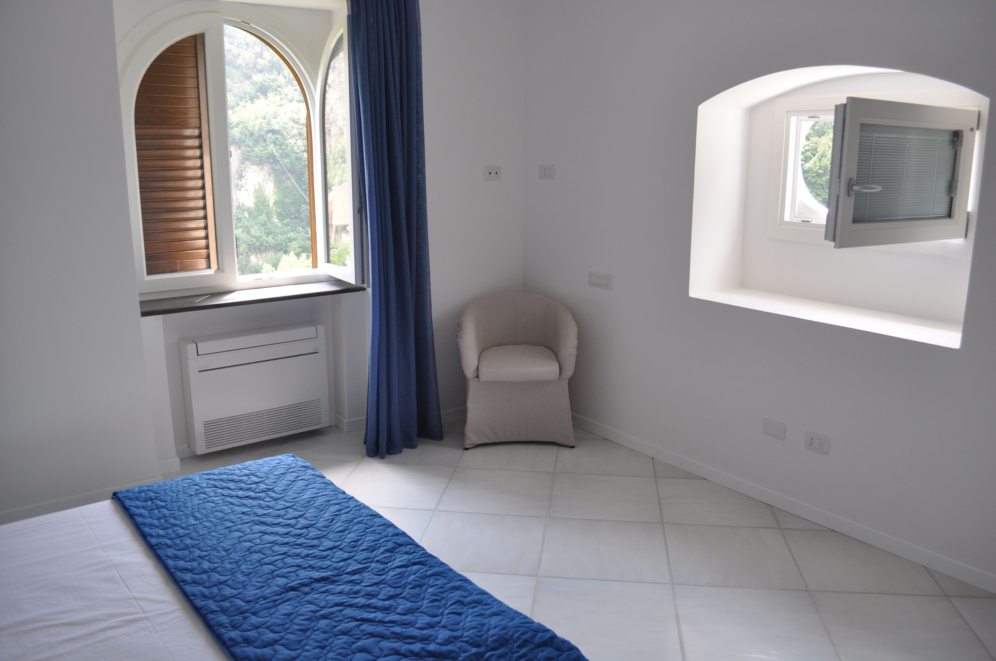 A bedroom with a bed, a chair, a window with blinds, and a small skylight.