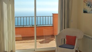 A balcony with a view of the sea, a wicker chair with a cushion, and a painting on the wall.