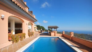 A swimming pool with a view of the sea, a covered outdoor seating area, and a building with an arched entrance.