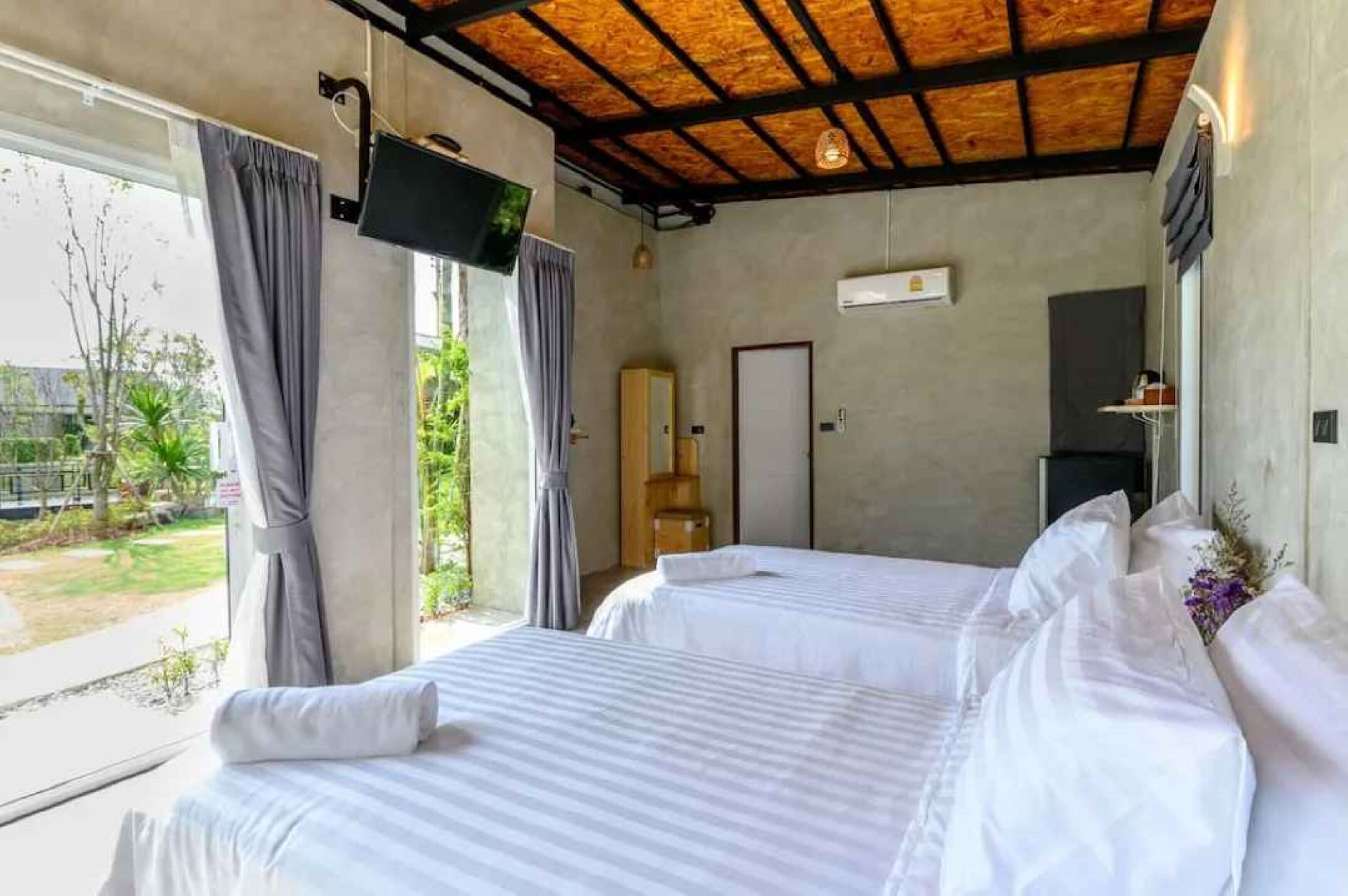 A hotel room with two beds, a TV, a window with curtains, and a view of greenery outside.