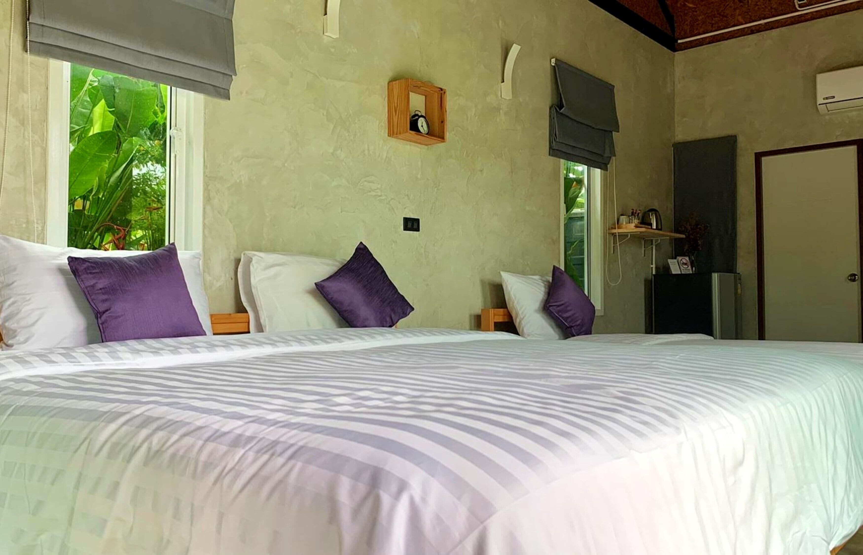 A bedroom with a large bed, two nightstands, a wooden headboard, a small shelf with a clock, and a window with a view of greenery.