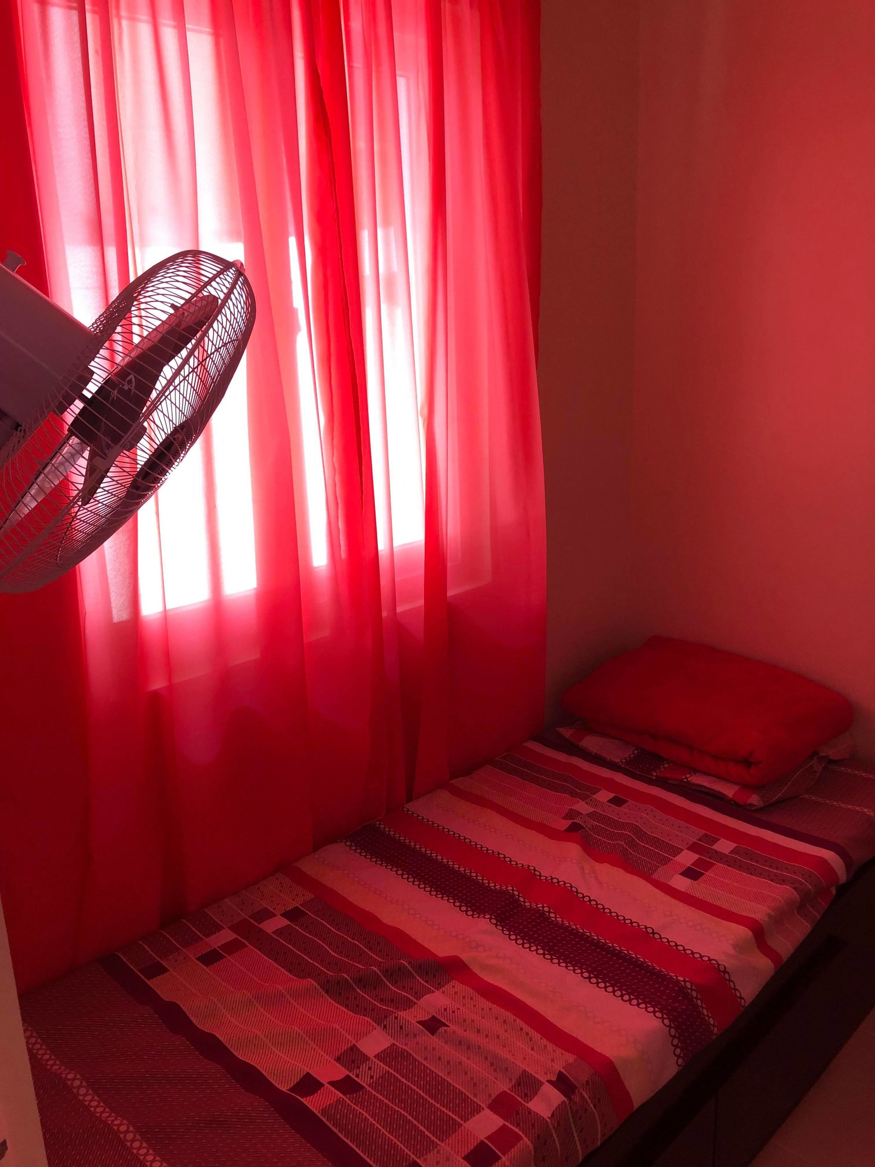 Una habitación pequeña con una cama, una cortina roja y un ventilador de pie.