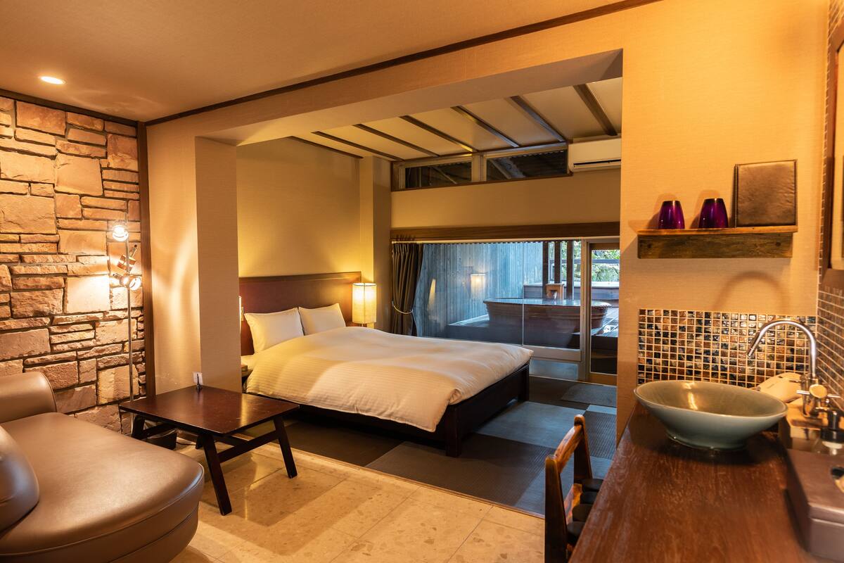 Ein Hotelzimmer mit Bett, Sitzecke, Steinwand, Badezimmer mit großem Waschbecken und Blick ins Freie.
