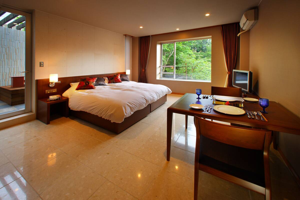 Ein Hotelzimmer mit einem großen Bett, einem Esstisch mit Stühlen, einem Fernseher und einem Fenster mit Vorhängen.