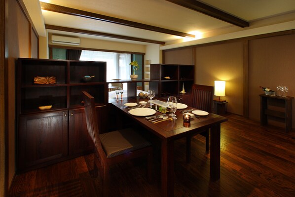 Un comedor con una mesa de madera dispuesta para cuatro personas, rodeada de sillas. Hay estanterías con objetos decorativos y una lámpara a un lado.