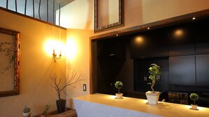 A reception area with a wooden counter, potted plants, a wall-mounted light fixture, and a dark-colored wall with a mirror. - Fujinokura Tiny Fort (Fujiyoshida)