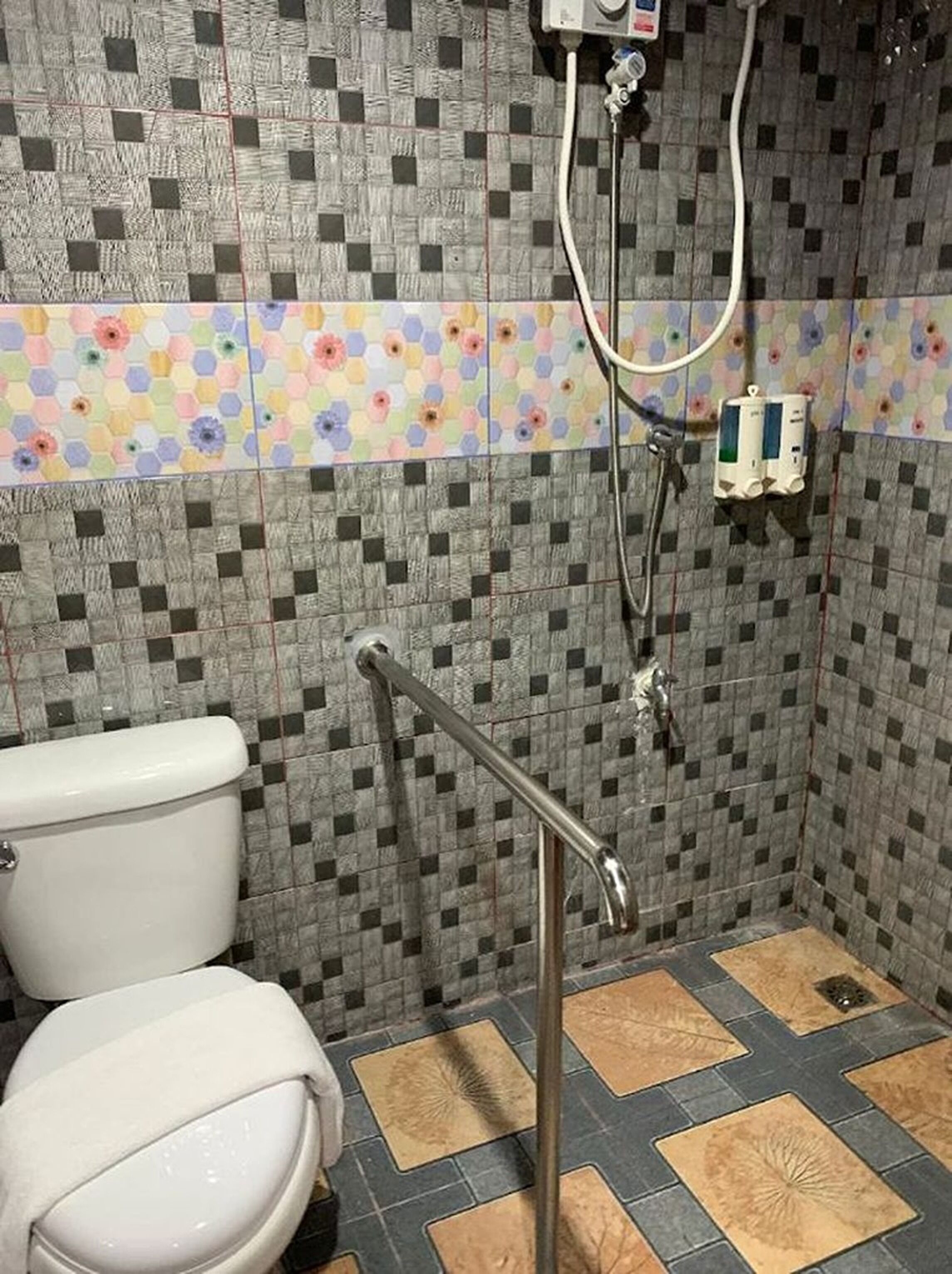 A bathroom with a toilet, a shower with a handrail, and a tiled wall with a floral pattern.