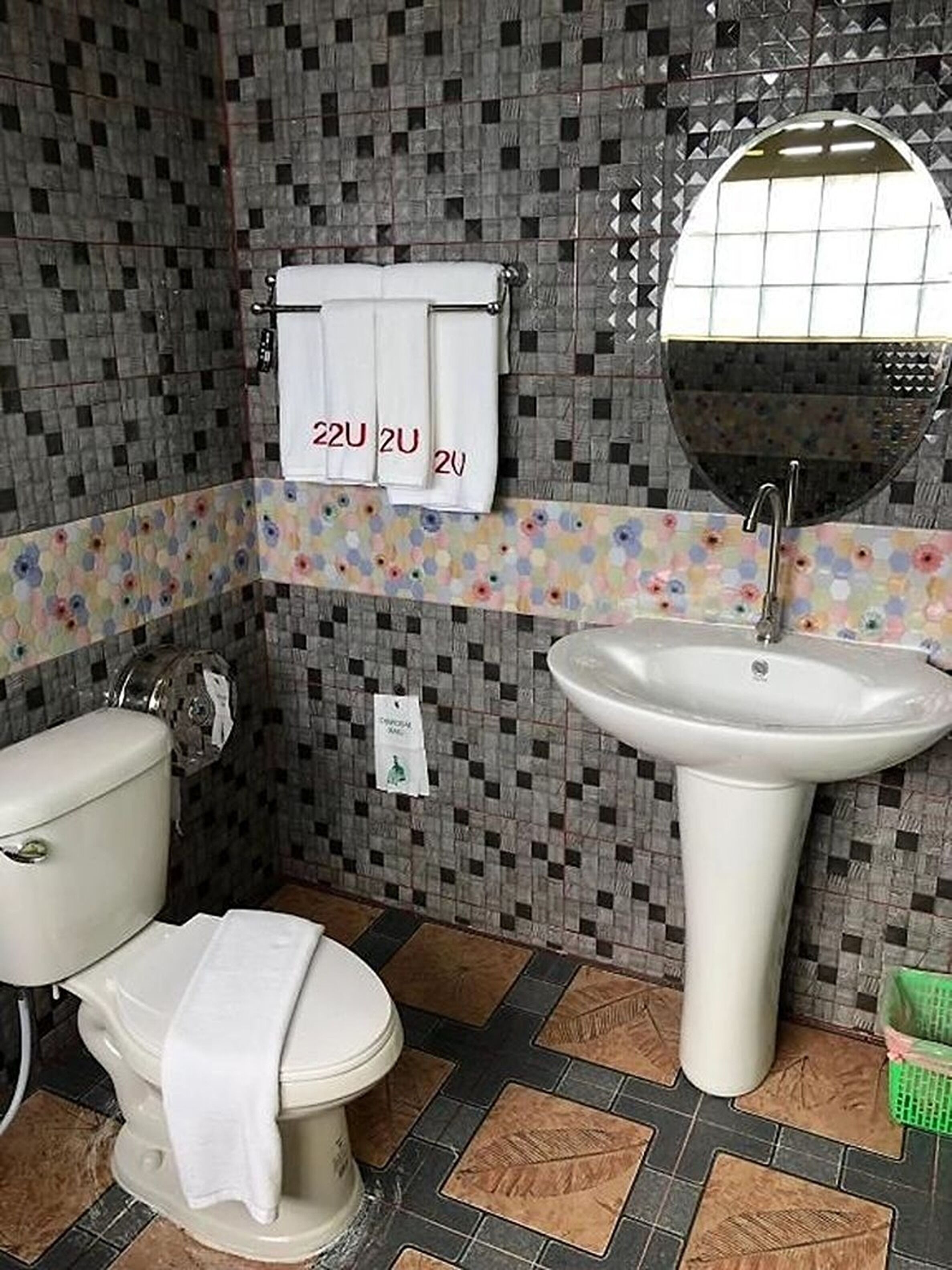 Baño con inodoro, lavamanos y espejo. Las paredes están cubiertas de baldosas con un estampado a cuadros, y hay un perchero con toallas.