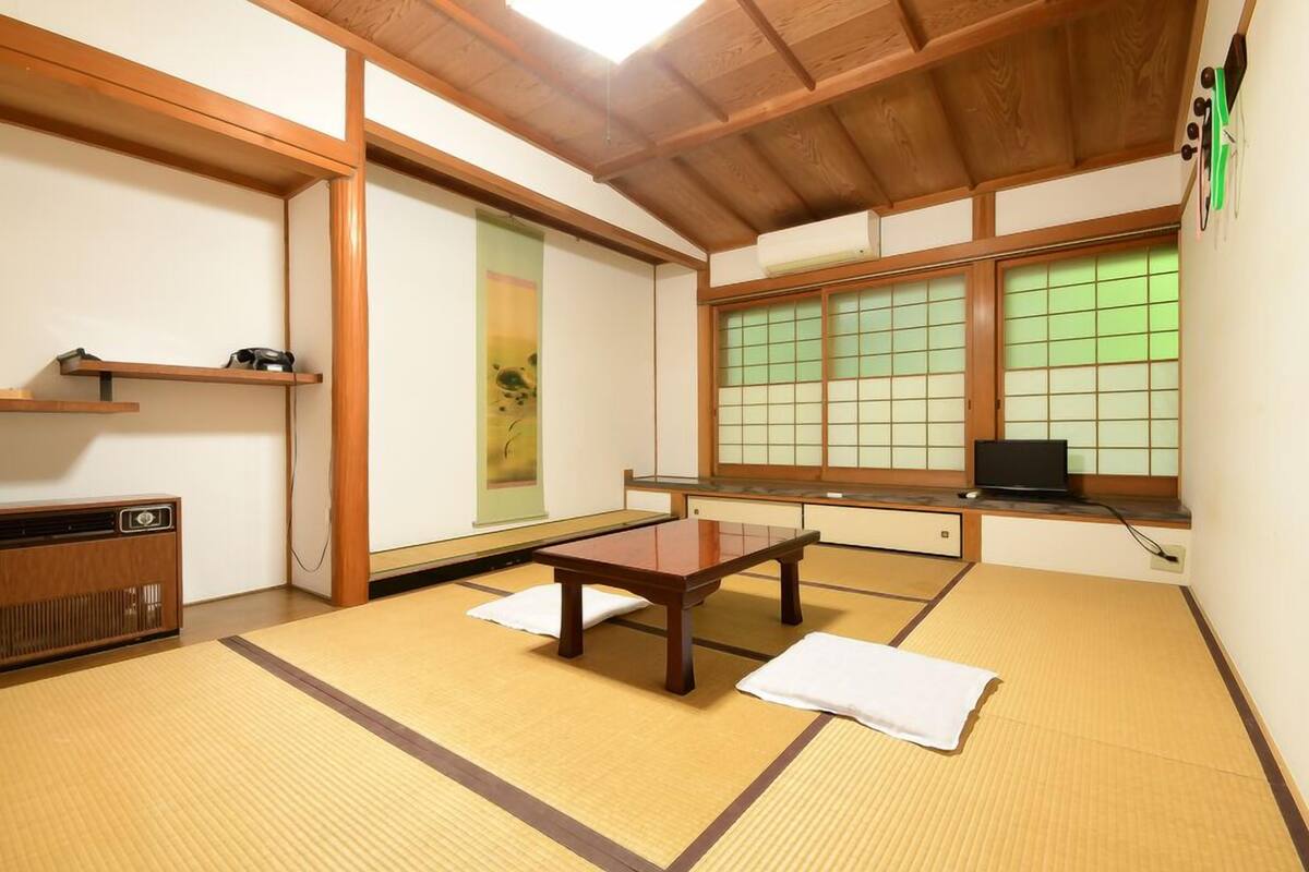 A tatami mat room with a wooden table, tatami mats, a radiator, a shelf with a book, a window with green glass, and a laptop on the window sill.