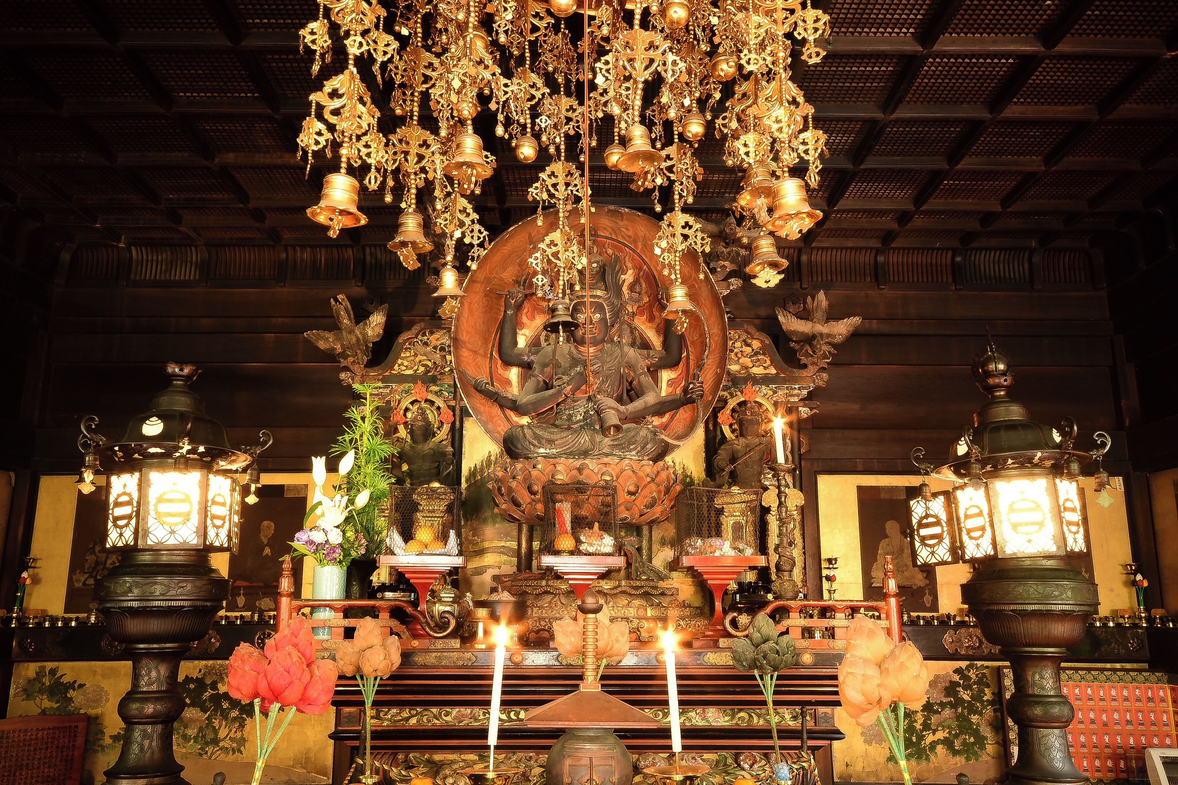 An ornate temple interior with a central deity statue, hanging bells, and traditional lamps.