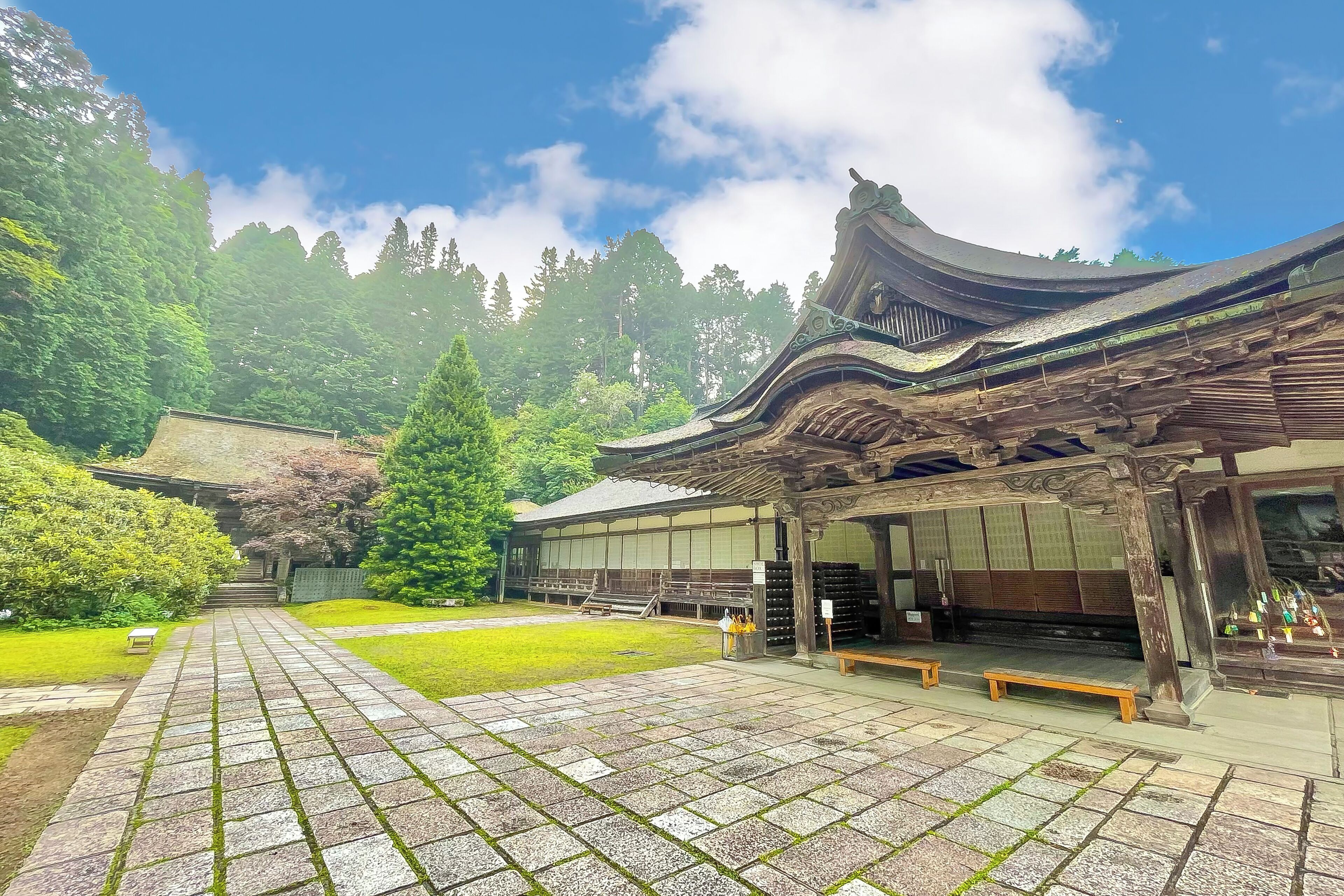 Un bâtiment traditionnel japonais, avec un chemin pavé menant à l’entrée, entouré de verdure et sous un ciel dégagé.