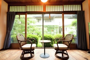 A room with two wicker chairs and a small round table, a large window with a view of greenery and a building, and wooden flooring. - Kongo Sanmaiin (Koya)