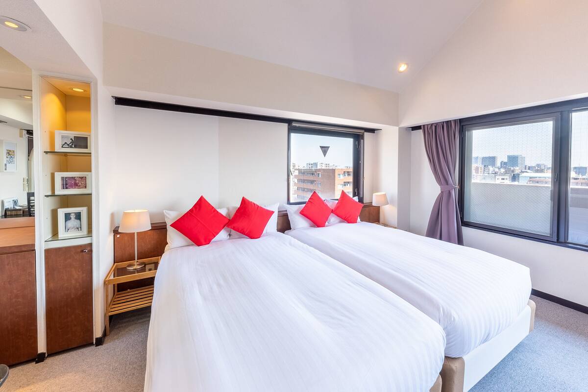 Une chambre d’hôtel avec deux lits, une grande fenêtre donnant sur la ville, une table de chevet avec une lampe et une étagère avec des photos encadrées.