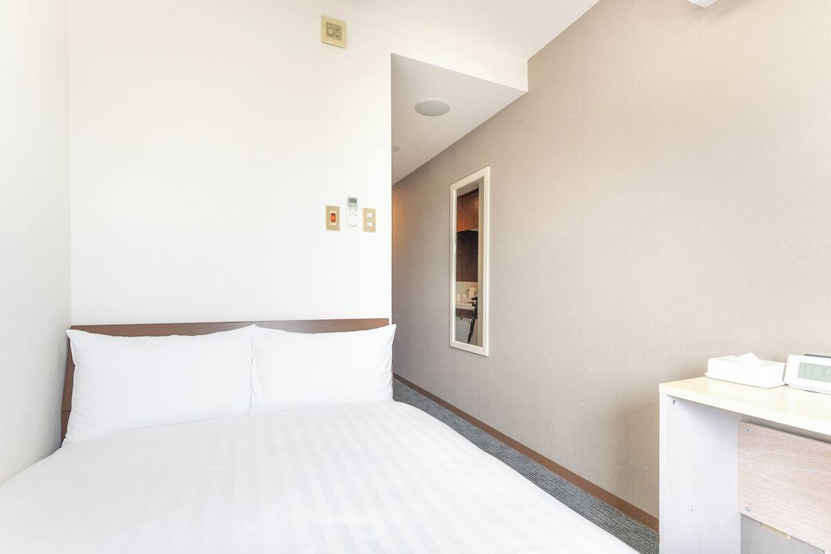 Une chambre d’hôtel avec un lit, un bureau et un miroir. Il y a deux interrupteurs sur le mur et un luminaire au plafond.