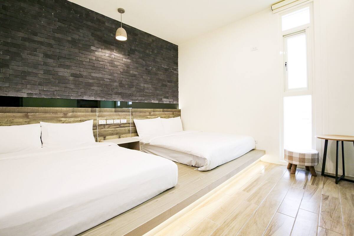 A modern hotel room with two beds, a brick accent wall, a wooden headboard, and a small table.