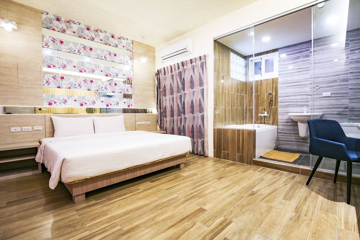 A modern hotel room with a large bed, a bathroom with a glass shower enclosure, and a blue chair.
